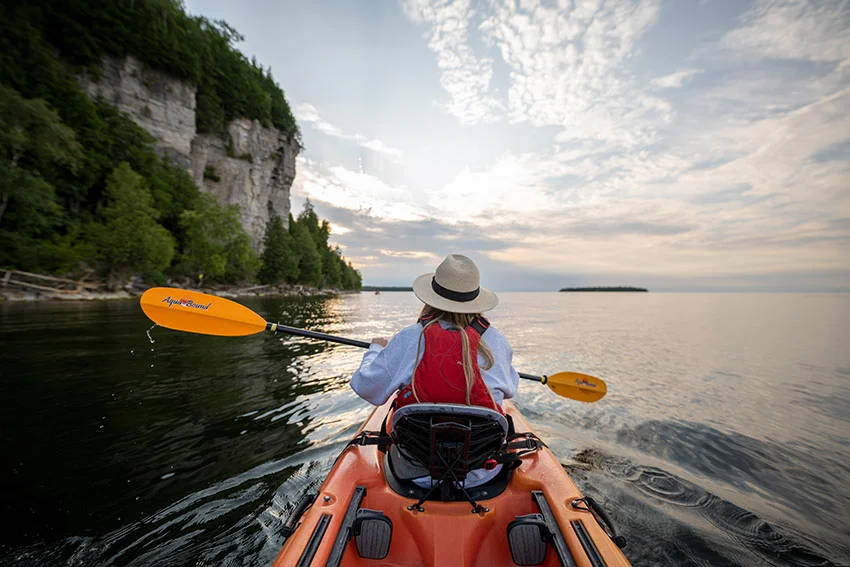 A kayaker paddling past the limestone bluffs and calm water of a Door County shoreline at golden hour