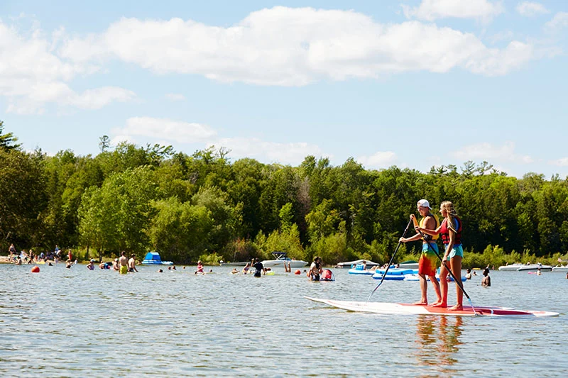 Two paddleboarders on a sunny Door County lake, with swimmers and boats along a tree-lined shore